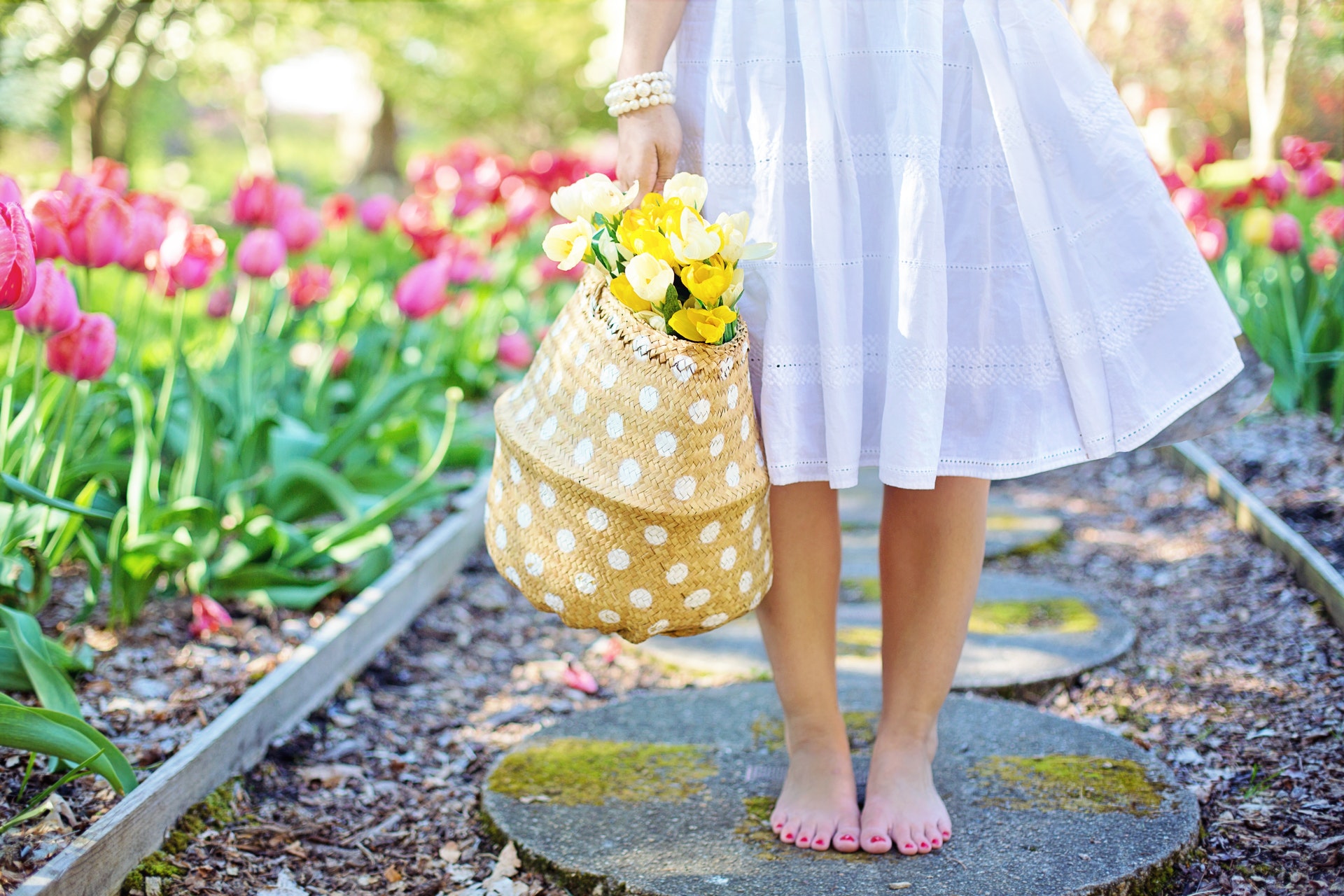 Lady in a white dress walking barefoot on a stone footpath with blooming flowers on the side.